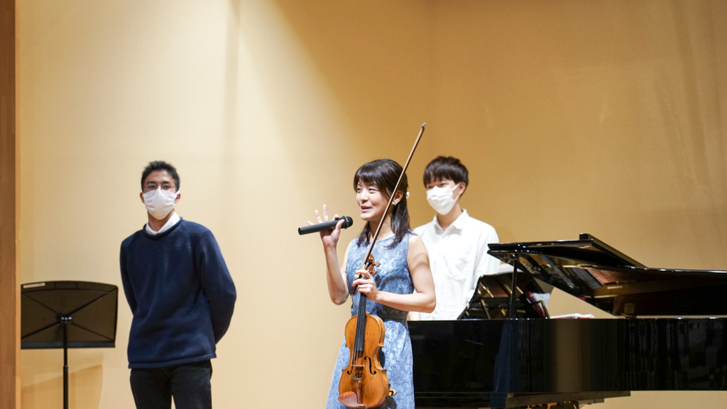 ヴァイオリンを片手に曲目紹介する廣津留先生の写真。傍らに立つのがピアノ伴奏をした仲田さん。